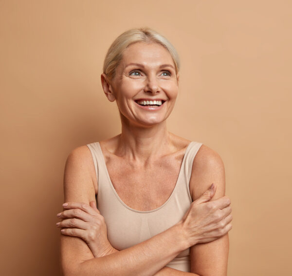 Attractive positive wrinkled fifty years old woman looks gladfully above keeps arms foded has well cared complexion healthy skin white teeth isolated over brown background. Beauty and age concept