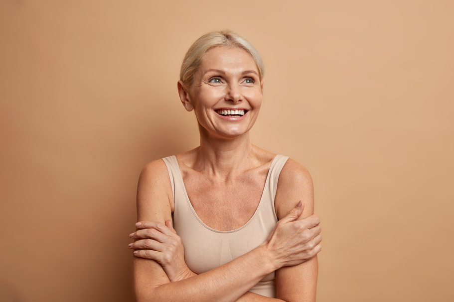 Attractive positive wrinkled fifty years old woman looks gladfully above keeps arms foded has well cared complexion healthy skin white teeth isolated over brown background. Beauty and age concept